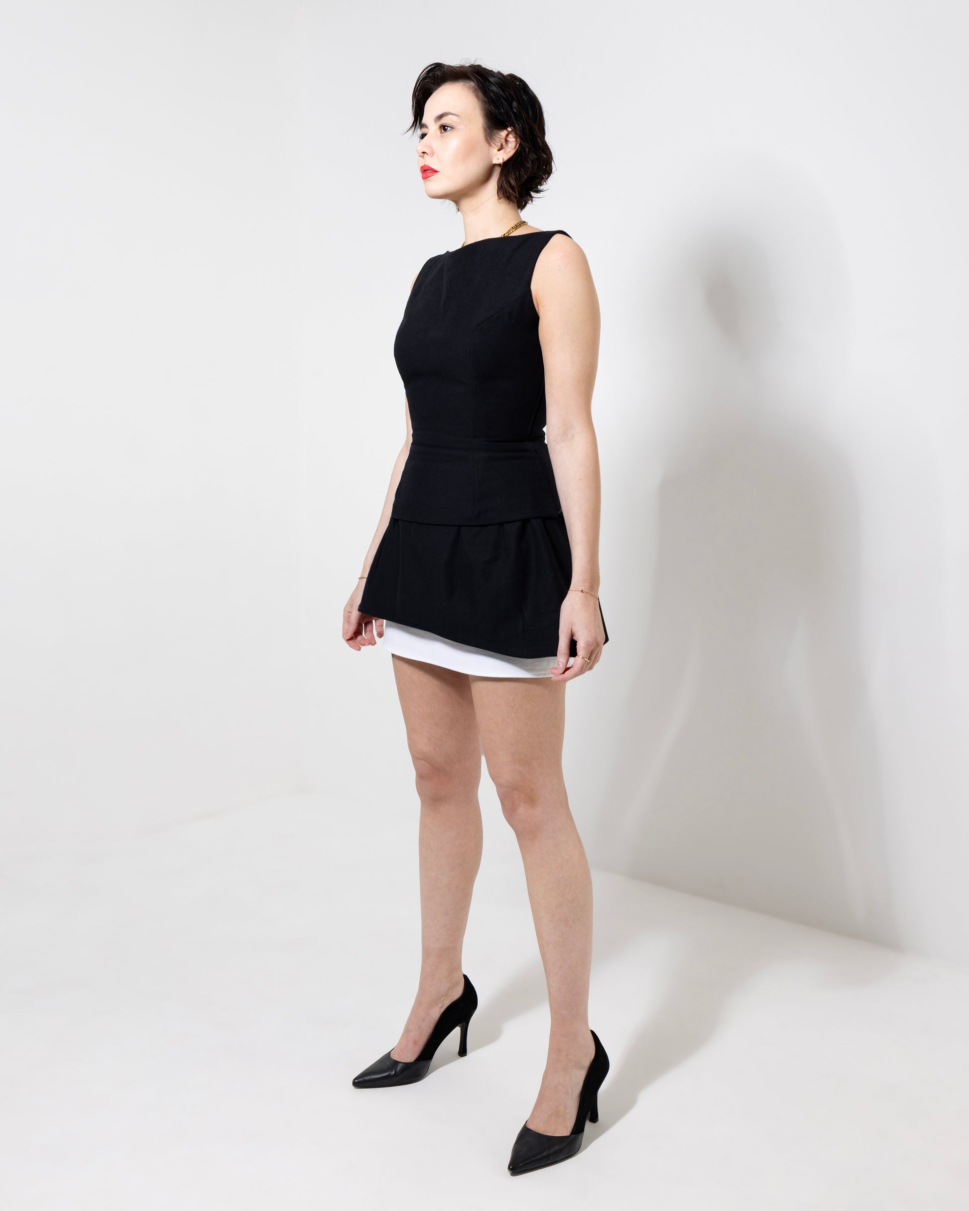 Model stands in front of white backdrop wearing Sans Seasons 'Lily' top in black - made from 100% cotton twill. A sleeveless boatneck top, upper hip length, with a cinched waist, hour glass shape, fastening with a zip up back - over the 'Calla' skirt. Model wears a size S and is a 27" waist, 33" bust and 5'8" tall